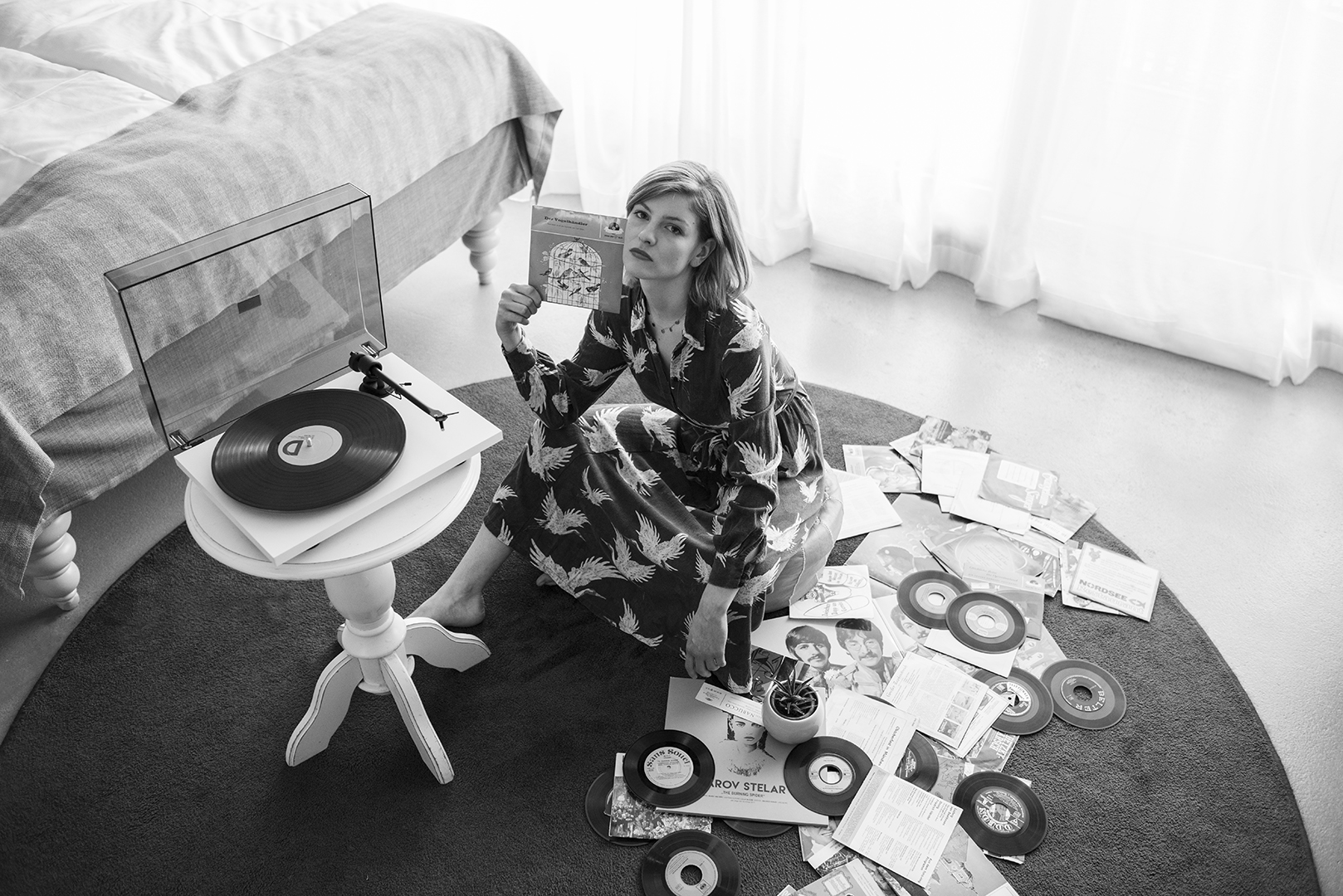 T1 Turntable sorounded by records on the floor and a woman sitting next to the turntable holding a record in the air. Ii is located in the bedroom